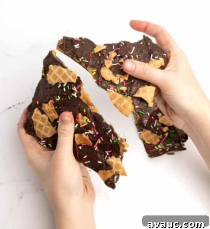 Christmas Nutty Buddy Bark 2 Hands gently breaking apart a sheet of homemade chocolate peanut butter bark with waffle cone pieces and colorful sprinkles, showcasing its crunchy layers.
