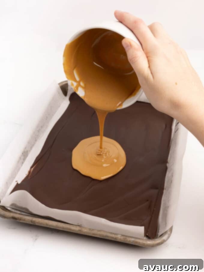 Christmas Nutty Buddy Bark 5 Melted peanut butter being poured from a small bowl onto a hardened dark chocolate layer in a parchment-lined baking pan.