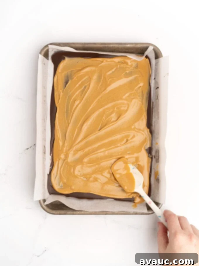 Christmas Nutty Buddy Bark 6 A spatula gently spreading melted peanut butter over the dark chocolate layer, ensuring an even and smooth coating for the bark.
