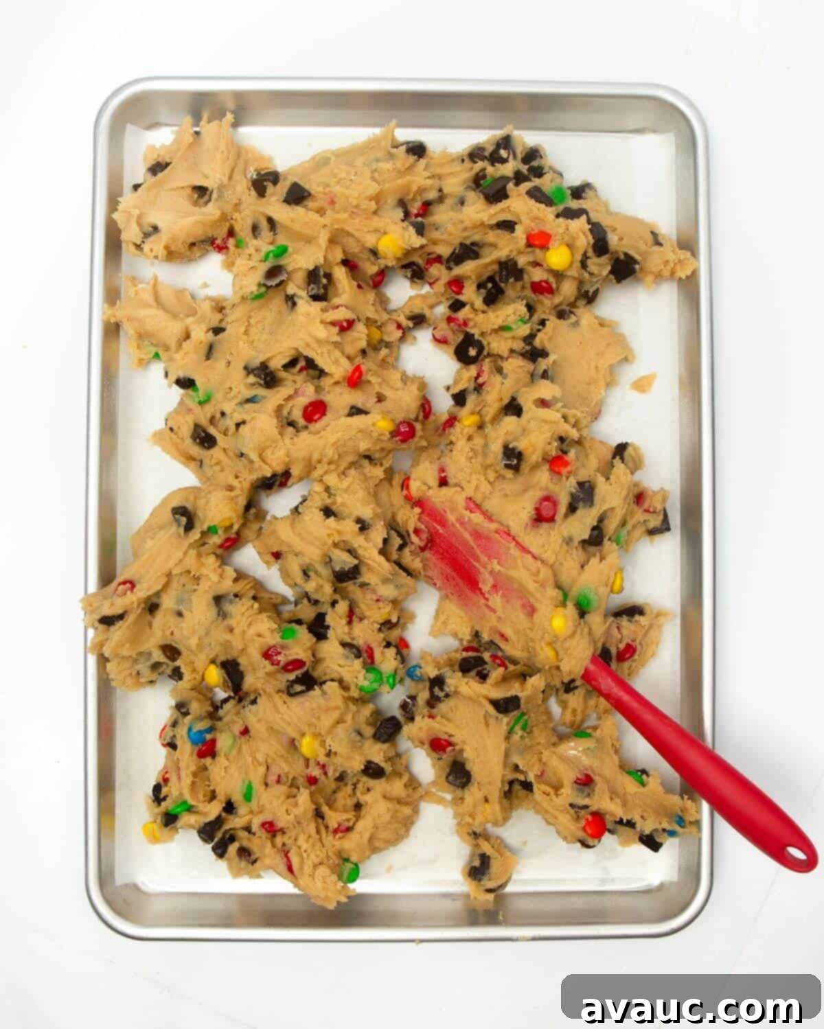 Spreading sticky cookie dough onto a parchment-lined metal pan with a spatula, ensuring an even layer.