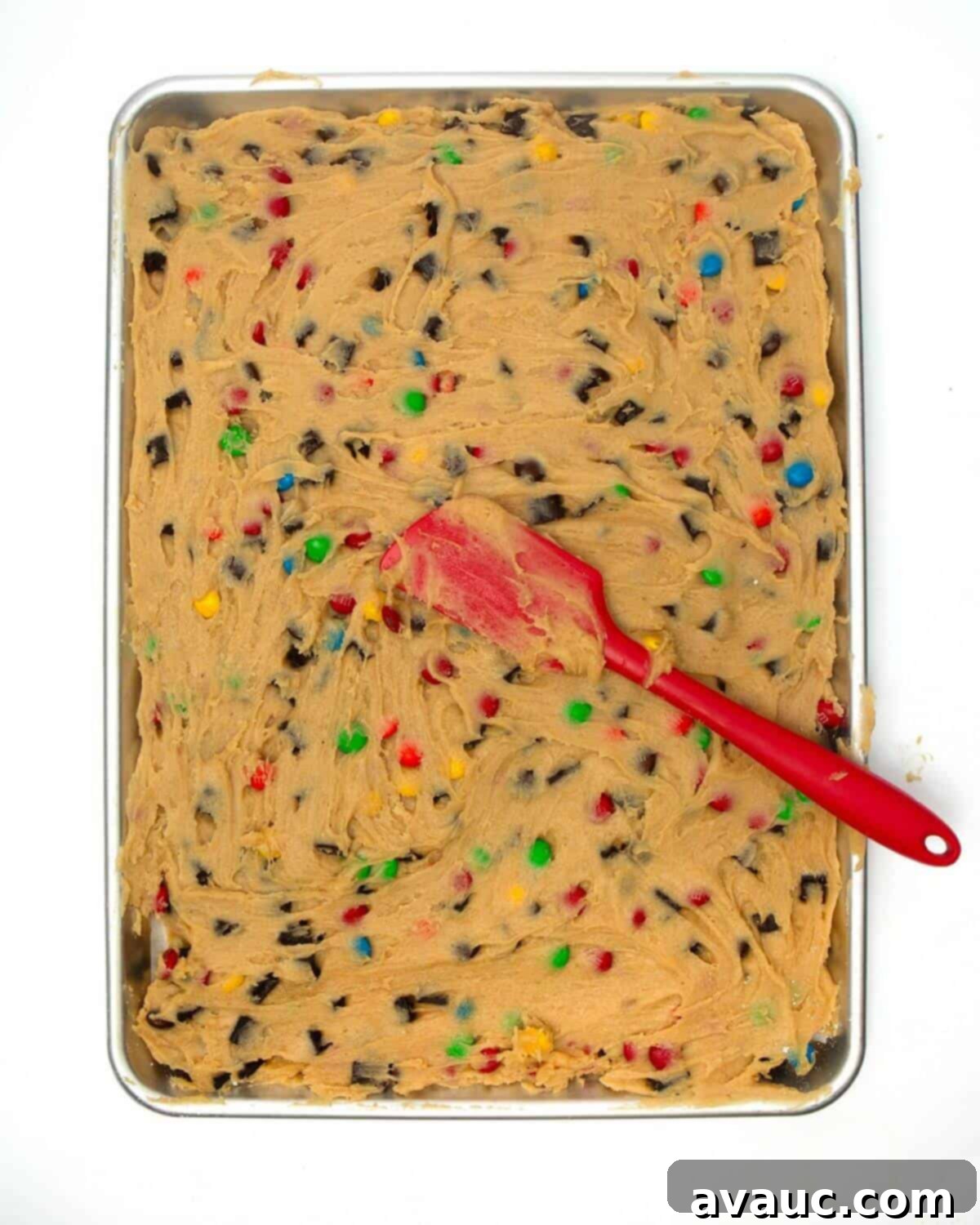 Cookie dough spread evenly across a metal pan with a spatula, ready for the next step of cutting.