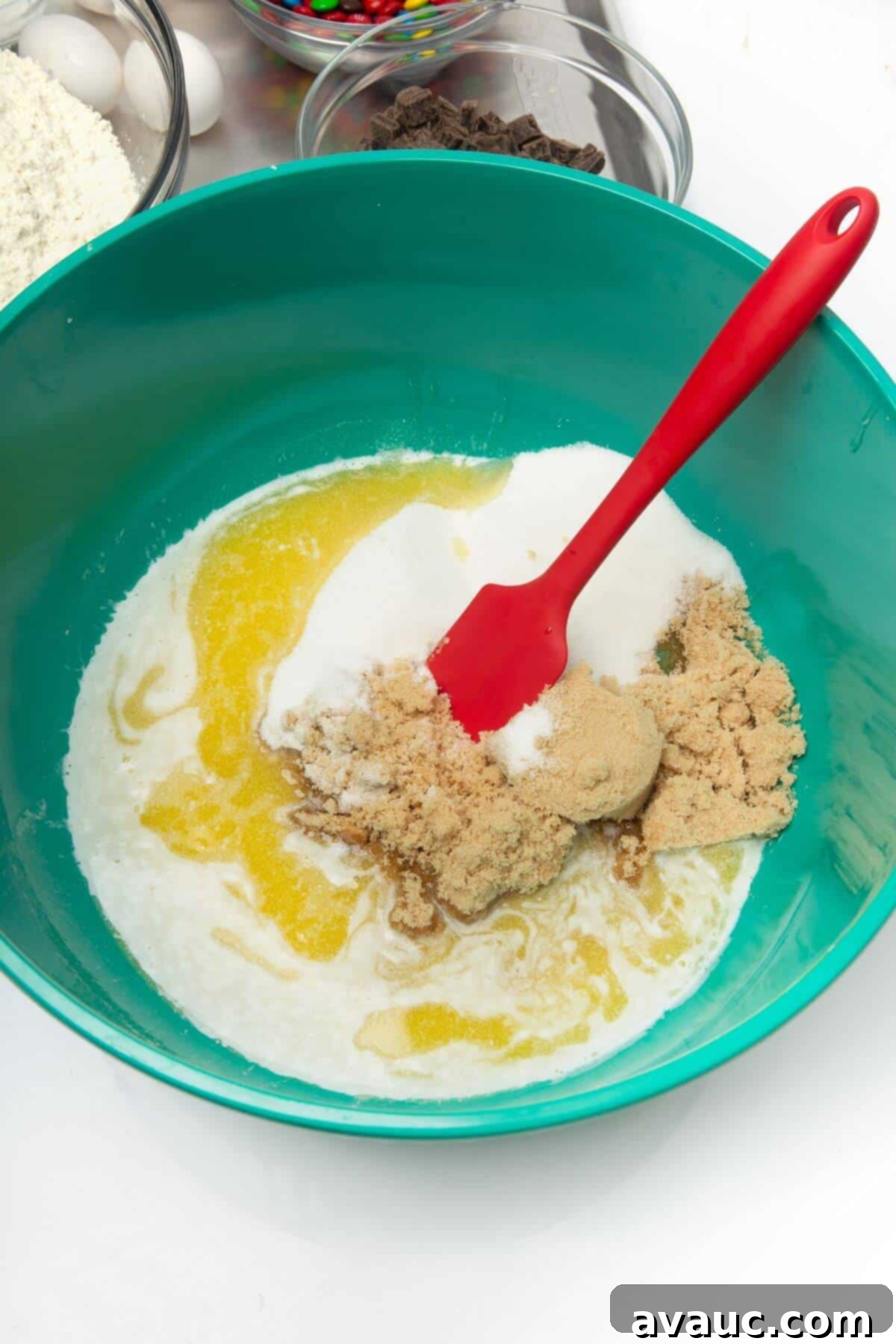 Light brown sugar and granulated sugar added to melted butter in a green mixing bowl, ready to be stirred with a red spatula.