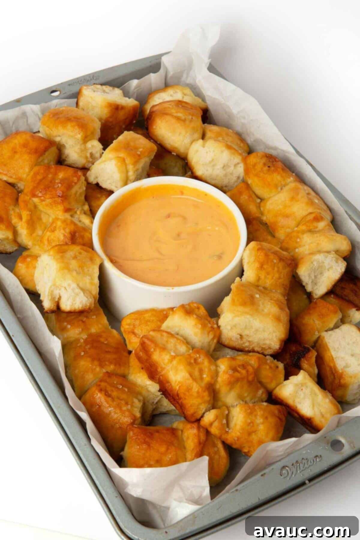 Golden brown pretzel bite monkey bread in a rectangular baking tray, with a side of creamy cheese dipping sauce.