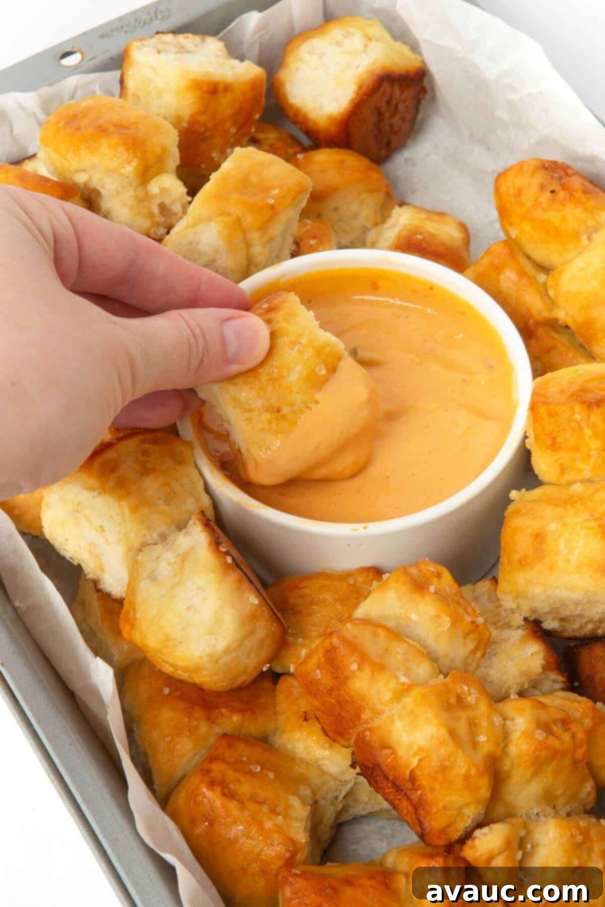 A hand dipping a piece of warm pretzel monkey bread into a bowl of creamy cheese sauce.