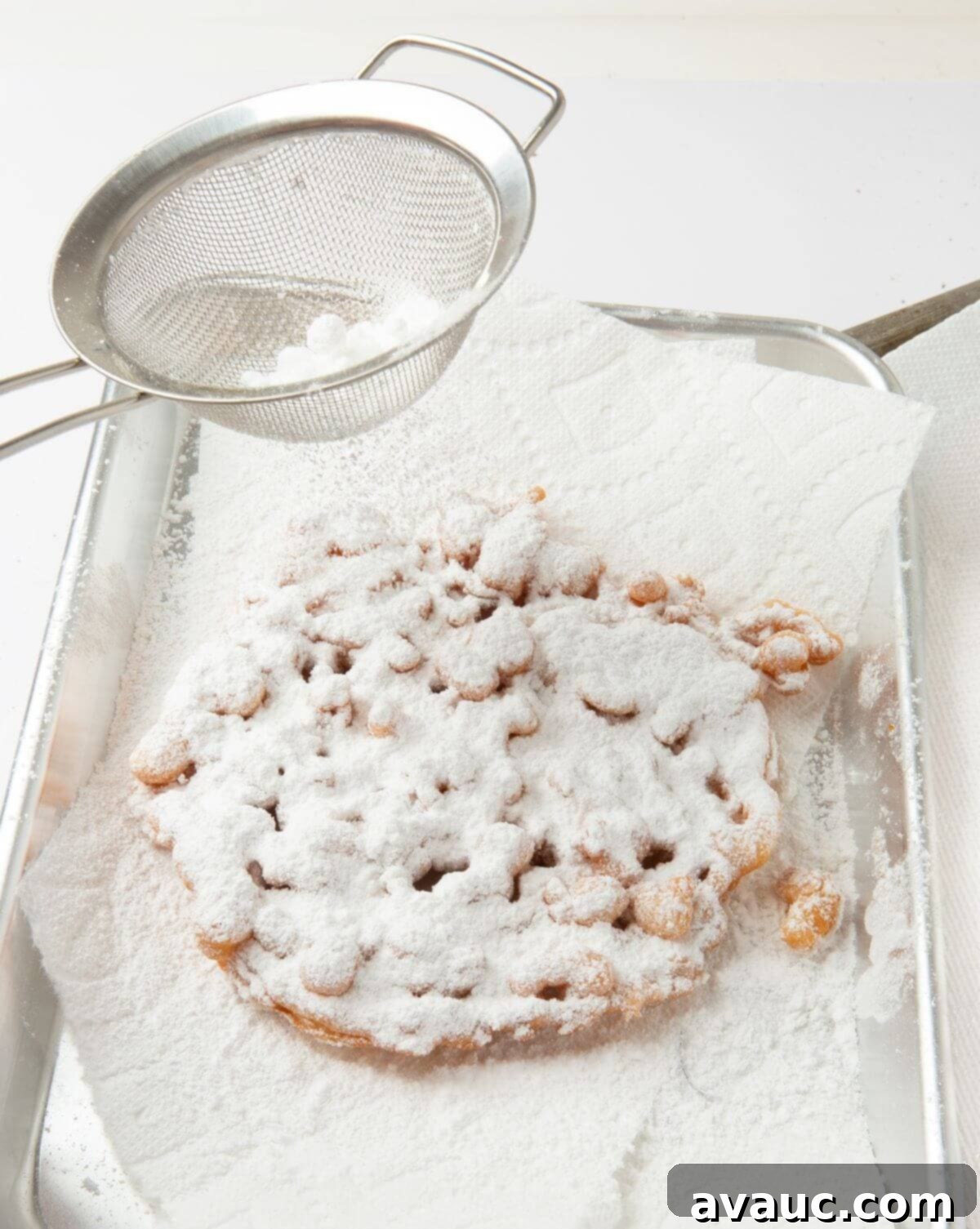 Simple Homemade Funnel Cakes 10 funnel cake on paper towel lined tray being sprinkled with powdered sugar using mesh colander