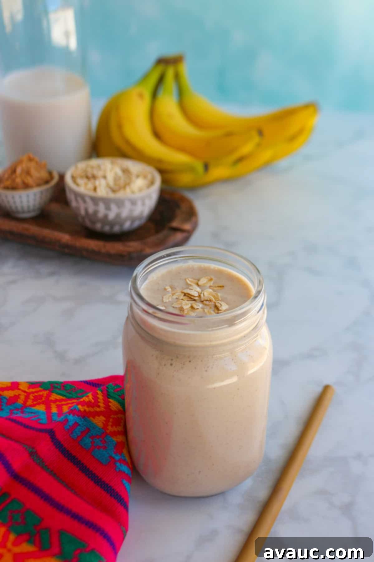 Glass filled with licuado de platano con avena (banana oatmeal smoothie) with a straw beside it and the ingredients behind it