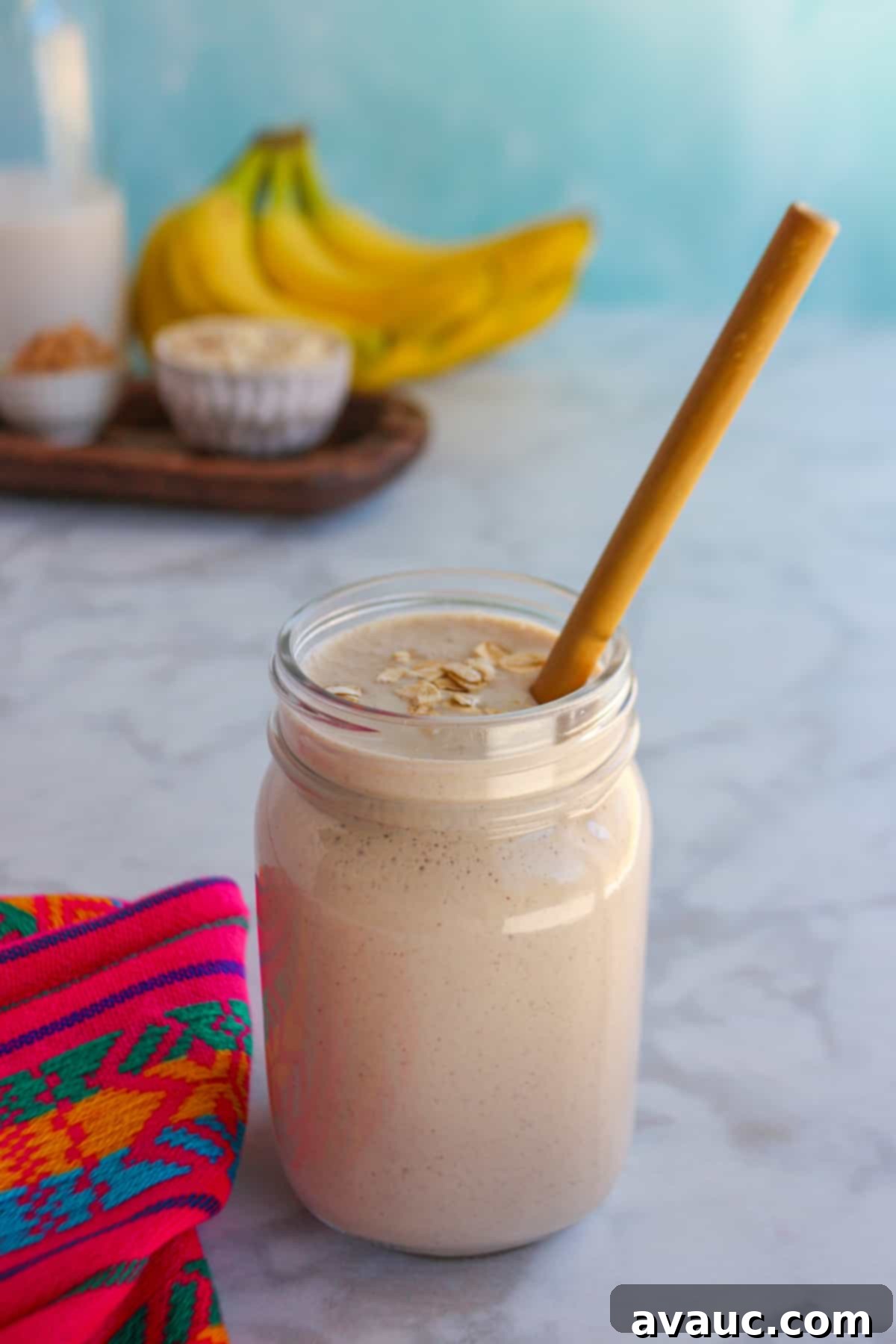 Glass filled with licuado de platano con avena (banana oatmeal smoothie) with a straw and the ingredients behind it