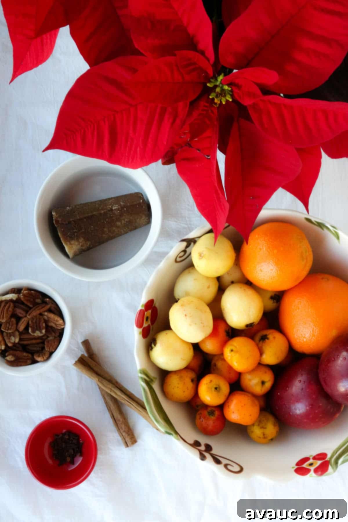 A poinsettia, a piloncillo cone, a small bowl with pecans, cinnamon sticks, a small bowl with whole cloves and a large bowl with oranges, guavas, tejocotes and apples