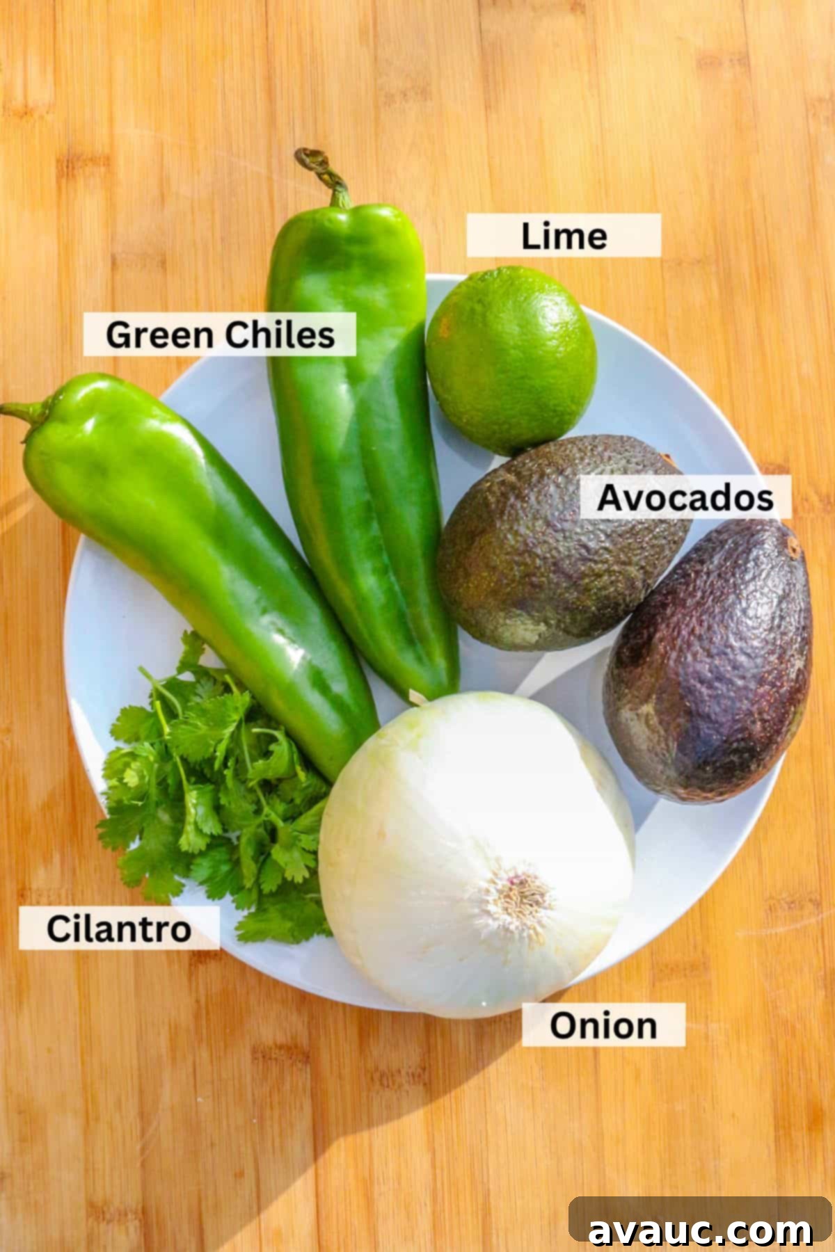 Smoky Roasted Green Chile Guacamole 3 A vibrant display of fresh ingredients for guacamole: green chiles, limes, ripe avocados, an onion, and a bunch of cilantro, arranged on a rustic wooden board.