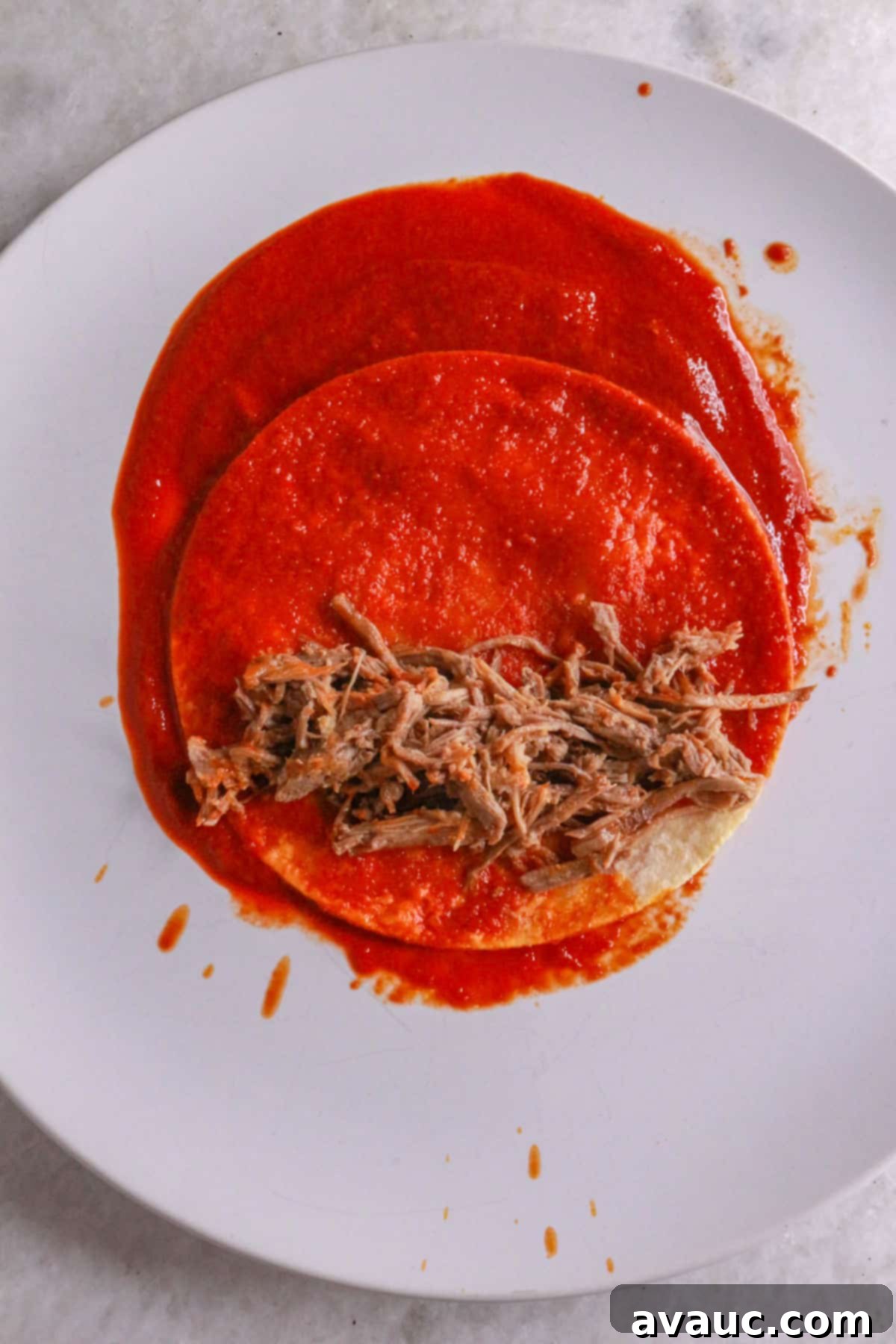 Classic Shredded Beef Enchiladas Rojas 9 Hands assembling an enchilada on a plate, adding shredded beef to a sauce-dipped tortilla before rolling.