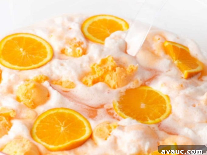 Creamy Orange Fizz 10 A glass of Orange Sherbet Punch with a decorative straw and orange slice, with a blurred background of a party setting.