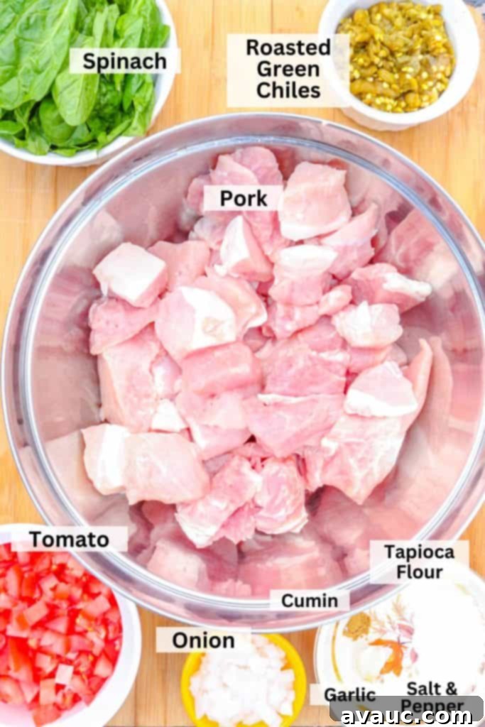 A vibrant display of fresh ingredients for pork green chile, including a bowl of raw, cubed pork surrounded by smaller bowls of spinach, fire-roasted green chiles, ripe tomatoes, diced onion, aromatic spices, and minced garlic.
