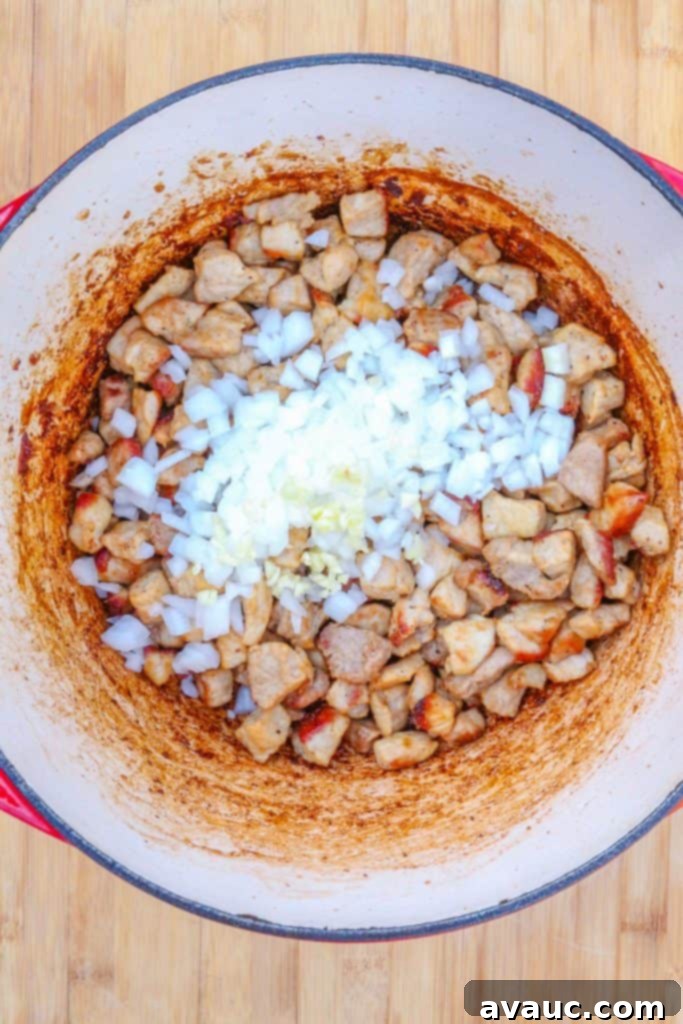 A Dutch oven with browned diced pork, now sautéing with fragrant garlic and diced onion, building a rich aromatic foundation.